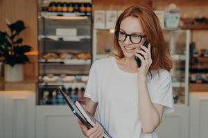 Happy ginger shop owner has telephone conversation holds mobile phone near ear photo