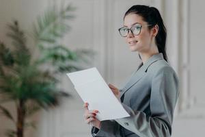 Successful brunette businesswoman stands thoughtful with paper documents, busy with making startup, analyzes business information, writes down notes, looks somewhere into distance, dressed formally photo