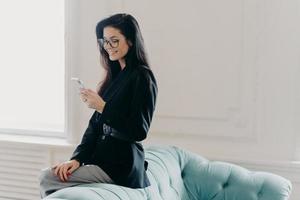 European entrepreneur wears corporate suit and spectacles, uses online banking application for transferring money, holds cellular, sends email, poses at back of comfortable sofa, uses free wifi photo
