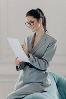 Vertical shot of successful female manager checks finance report, involved in working process, signs inportant contract, has happy expression, wears formal suit, poses in living room, works from home photo