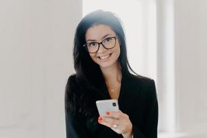 Happy business consultant dressed in formal wear, holds mobile phone, sends text messages, reads news online, poses indoor at office, has hard working day. People, technology, entrepreneurship concept photo