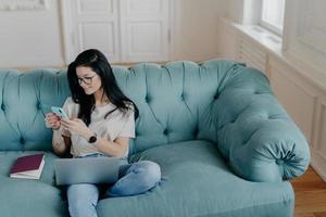 View from above of busy female freelancer sits in cozy room on comfortable sofa, works on new startup project, uses laptop computer, holds mobile phoneand sends text messages, interacts with netbook photo