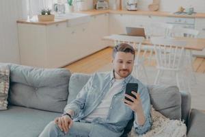 Attractive caucasian young man in domestic casual clothes using smartphone at home photo