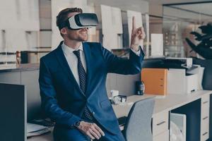 Enjoyed young businessman pointing with forefinger up in air with virtual reality glasses on head photo