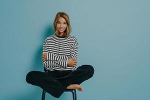 Relaxed young girl in striped shirt sitting with legs folded together in lotus pose on top of chair photo