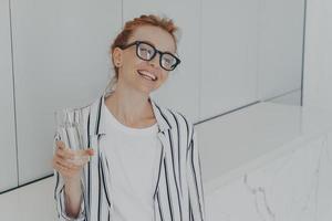 Young beautiful pleased red-haired woman having healthy habits, drinking mineral water in morning photo