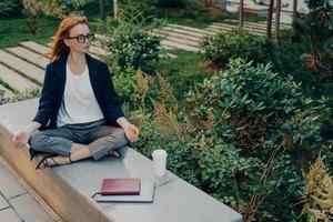 Relaxed ginger woman meditates in park has peaceful meditation enjoys nature