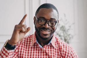 un tutor de inglés masculino de etnia africana sonriente con gafas levanta la mano y señala con el dedo índice hacia arriba foto