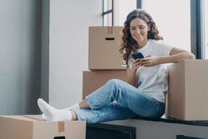 Happy spanish woman is sitting on windowsill texting on phone and unpacking boxes. photo