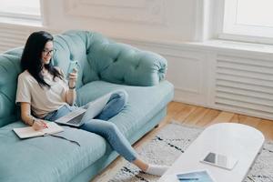 Above shot of pretty brunette woman focused in samrtphone display, writes down necessary information in notebook, watches training webinar, poses in cozy room on comfortable couch, works from home photo