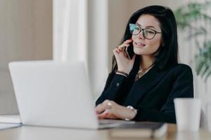 Confident serious business lady has phone conversation, poses in front of laptop computer, wears optical glasses for vision correction, poses at work place, makes reserach work, dressed formally photo