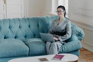 Horizontal shot of contemporary successful businesswoman poses in spacious room on comfortable sofa, reads useful article for business development, works on laptop computer, wears elegant clothes photo