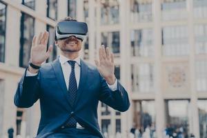 Young happy bearded man office worker dressed formally sitting outdoors and using VR headset or 3d goggles photo