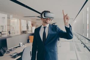 Happy man office worker wearing vr goggles, pointing with finger at objects in digital world photo