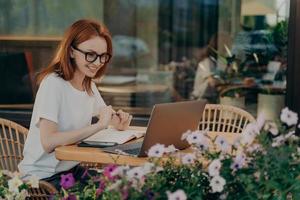 Redhead skillful woman student writes in notebook sits in front of opened laptop computer photo