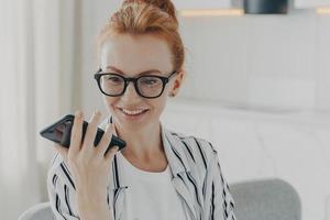 Beautiful positive red-haired woman in spectacles holding mobile phone and sending audio message photo