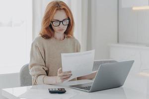 Young focused red-haired woman sitting at table with laptop and holding paper taxes bills photo