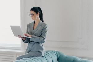 Photo of brunette executive worker poses in home office near sofa, holds laptop computer, makes financial report, wears optical glasses for vision correction, grey elegant suit, surfs internet
