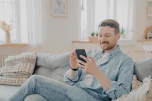 hombre barbudo sonriente feliz usando un dispositivo de teléfono inteligente mientras se relaja en el sofá foto