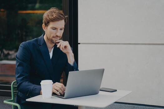 Handsome pensive young man browsing web on laptop while working remotely in sidewalk cafe photo