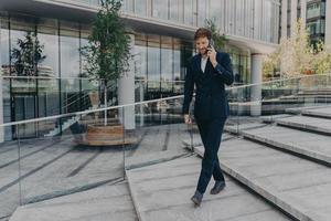 Happy caucasian businessman talking on smartphone while walking along office center photo