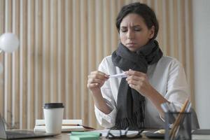 Unhealthy young hispanic business woman wearing scarf around neck holding thermometer photo