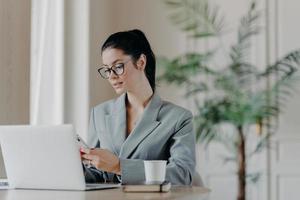 Gorgeous busineswoman types message on smartphone, works on new publication, poses in front of laptop computer, involved in working process, sits at desktop, drinks aromatic coffee, uses internet photo