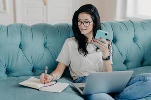 toma interior de una bella estudiante morena que ve un seminario web y toma notas en el bloc de notas, sostiene un teléfono móvil, se sienta en un cómodo sofá, ocupada con un trabajo a distancia. gente, tecnología, concepto de estilo de vida foto