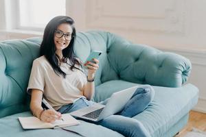 Glad female student works on course paper, surrounded with modern gadget, makes notes in notepad, feels relaxed at comfortable sofa, works online, wears casual clothes, watches video online. photo