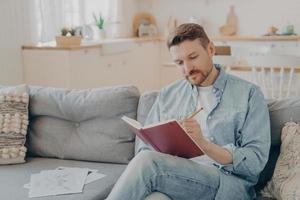 Busy young man writing in red notebook with pencil photo