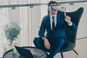 Smiling businessman using VR headset glasses while sitting in office lobby and working on laptop photo