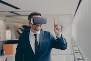 Smiling bearded office employee in suit wearing in VR glasses working in augmented reality in office photo