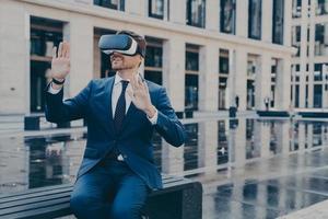 Man in VR goggles trying to reach something in virtual reality with his hands while sitting outdoors photo