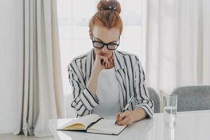 Redhead woman in striped shirt takes notes writes down information in notebook photo