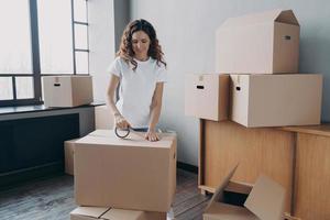 Young hispanic woman is packing containers with sticky tape. Happy mover wrapping cardboard boxes. photo