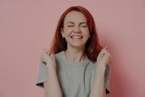 hopeful red-haired happy female making wish with closed eyes, isolated on pink background photo