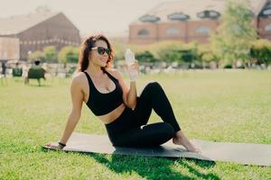 Happy young sporty European woman has rest after training poses on fitness mat with bare feet drinks water dressed in activewear sunglasses leads healthy lifestyle. People workout hydration concept photo
