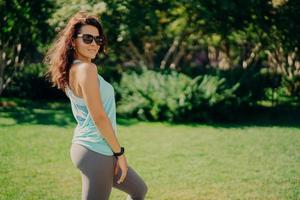 Pleased brunette European woman in active wear poses sideways wears sunglases smartwatch has fitness outdoors rests after jogging smiles pleasantly. Health body care sport and exercising concept photo