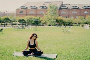 Relaxed slim brunette Caucasian woman sits in lotus pose on fitness mat has perfect body shape does yoga and meditation wears sports clothes poses outdoor. Health fitness and relaxation concept photo