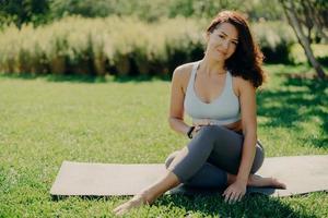 Photo of brunette sportswoman has rest after fitness training sits on karemat with bare feet in activewear leads healthy lifestyle poses on fresh green grass enjoys summy day and good weather