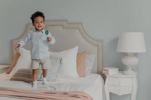 Carefree cute afro american child boy with lollipop jumping ob bed at home photo