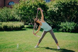 una foto completa de una mujer activa estira los brazos con goma de mascar tiene buena flexibilidad usa polainas de camiseta y zapatillas de deporte tiene entrenamiento con poses de banda de resistencia al aire libre en el césped verde durante el verano