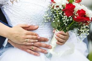 el novio abraza a la novia con un ramo de rosas rojas y blancas de boda con lirio de los valles. las manos con anillos de los recién casados foto