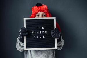 Boy wearing winter suit holding black sign saying it's winter time for winter concept photo