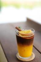 Iced Black coffee mixed with orange juice in glass topping with sliced oranges, rosemary leaves and cinnamon on top. on a wooden coaster placed on a wooden table.