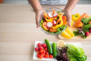 vista superior de una ensalada de verduras que se sostiene en la mano contra un fondo borroso de verduras en la mesa como tomates, pepinos, roble verde, roble rojo, limón en la cocina. foto