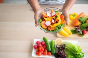 close-up top view of a vegetable salad holding in hand against a blurred background of vegetables on the table such as tomatoes, cucumbers, green oak, red oak, lemon in the kitchen. photo