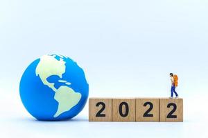 2022 New Year and Travel Concept. Closeup of woman traveler miniature figures with backpack standing on stack of wooden number block with mini world ball on white background. photo