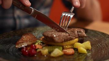 Mans hand holding knife and a fork cutting grilled beef steak on stoned plate. Selective focus video
