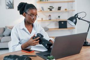 Holding camera. Young african american woman in white shirt is at home photo